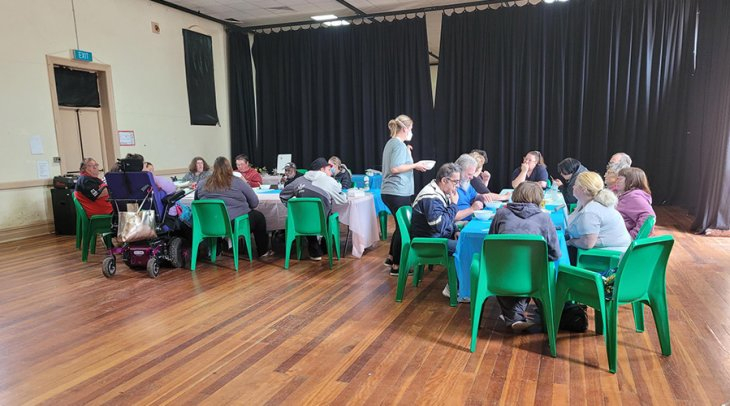 Numerous NDIS participants sitting around tables in a hall eating.