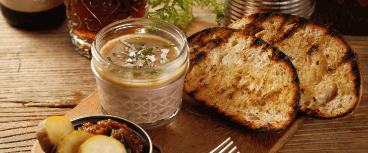 Two slices of toasted bread, with a jar of pate and some pickled gherkins and chutney